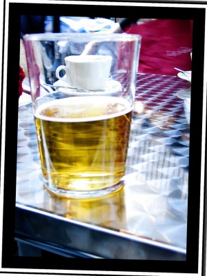 Cerveza sobre la mesa metálica en vaso de cristal a través del cual se ve el fondo un taza de café blanca